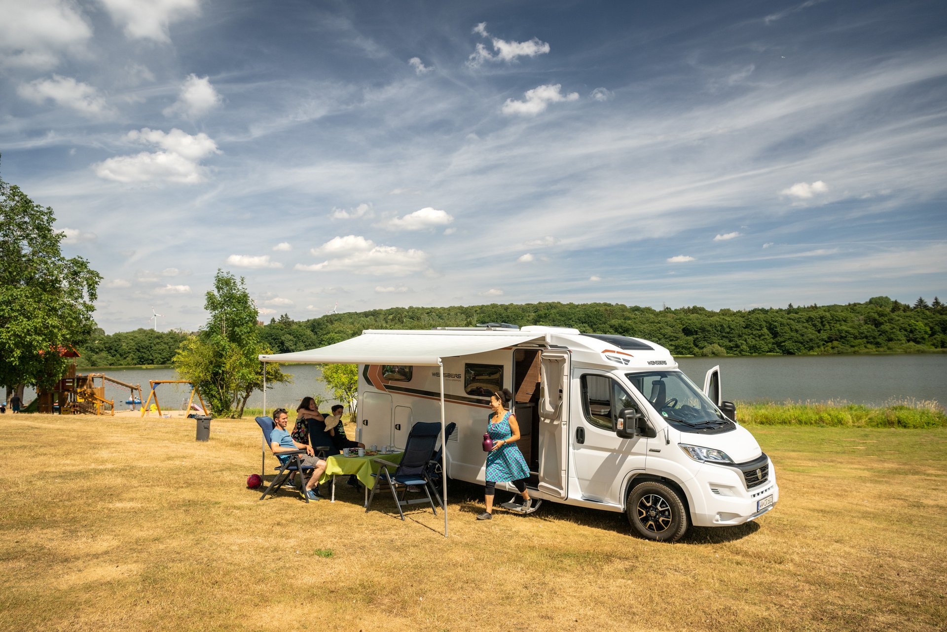 Familie sitzt unter der ausgefahrenen Markise Ihres Campers am Dreifelder Weiher