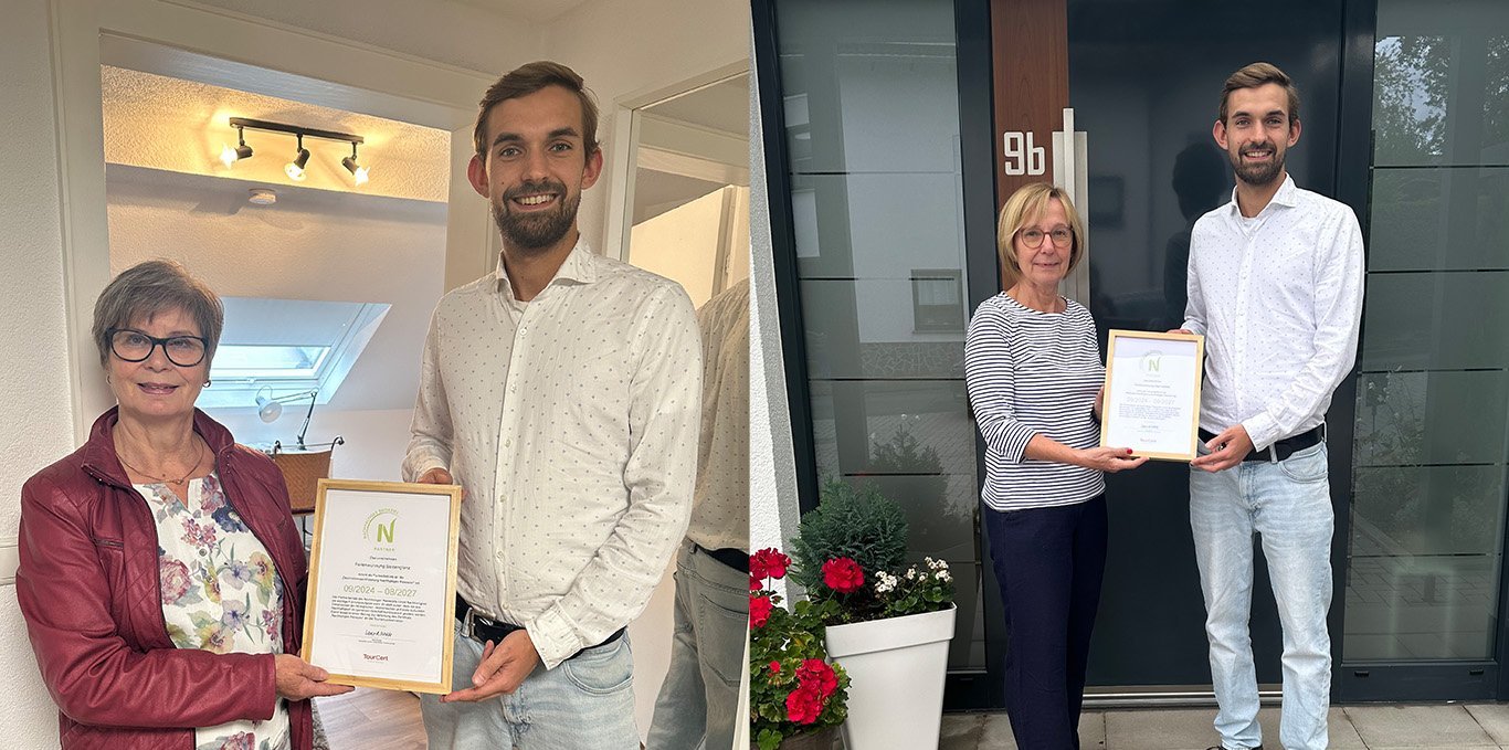 Zwei Personen halten Urkunden in der Hand, stehen vor einem Gebäude.