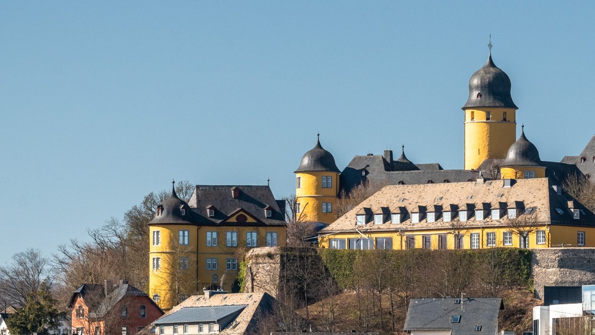 Eine Burg thront über einer Stadt mit gewöhnlicher Architektur im Vordergrund.